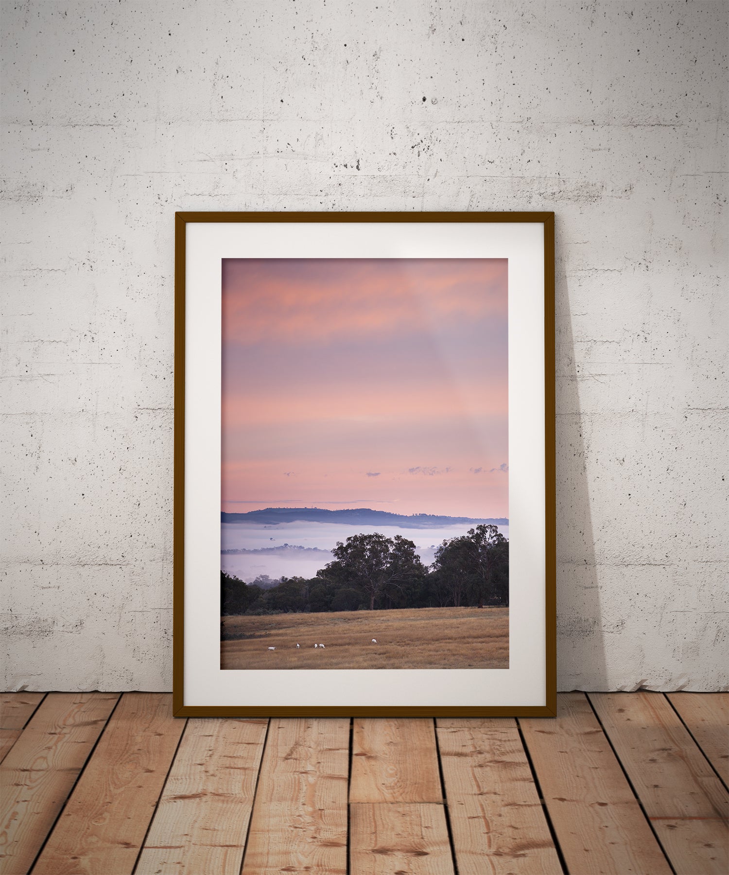 Framed artwork of a landscape with a pink sky hanging on a white brick wall.